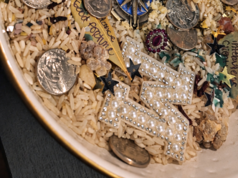 Close-up of a money bowl filled with rice, coins, jewelry, crystals, and handwritten prosperity petitions, with the title “Use What You Have: The Art of Substitution in Spellwork.”